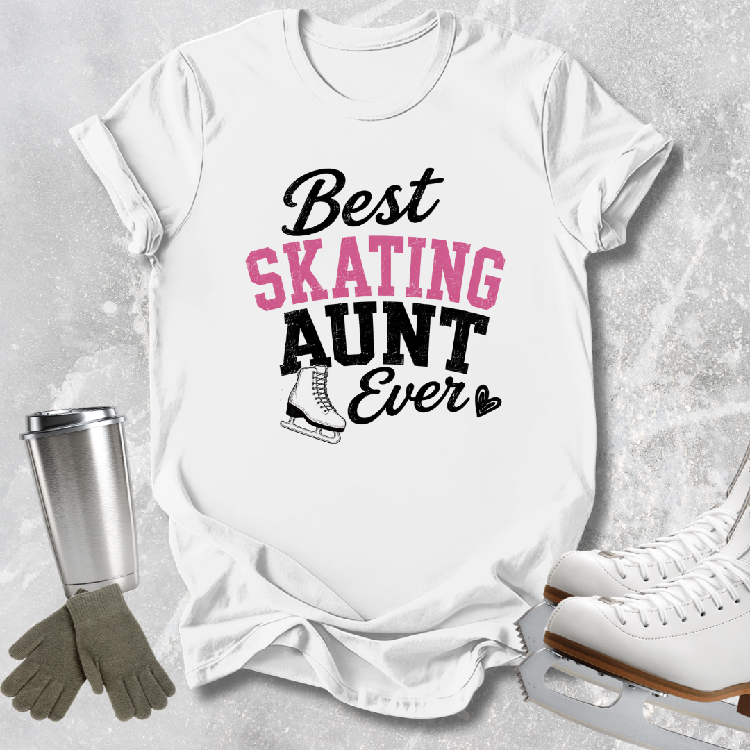A white graphic T-shirt laid flat on a light gray textured background, with the text 'Best SKATING AUNT Ever' in black and pink lettering and a small ice skate illustration. Surrounding props include a metal tumbler, gloves, and ice skates.