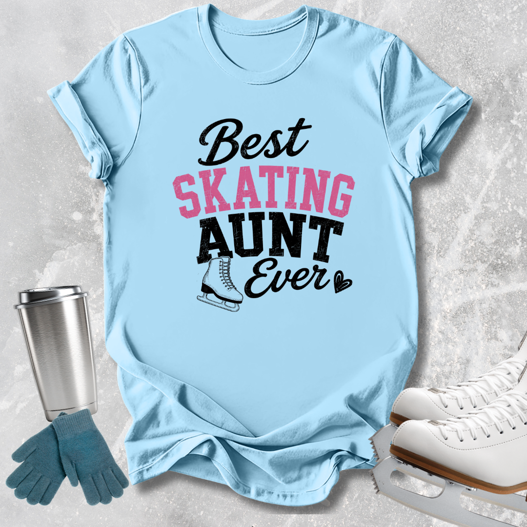 A light blue graphic T-shirt laid flat on a textured light gray surface, with a silver tumbler, teal gloves, and white ice skates around it. The shirt reads 'Best SKATING AUNT Ever' in black and pink lettering with a small ice skate illustration.