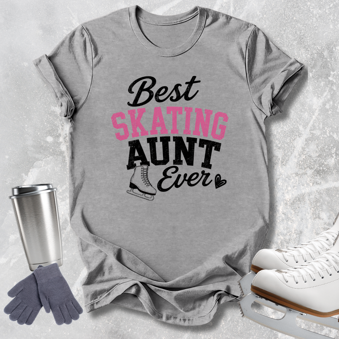 A heather gray graphic T-shirt laid flat on a light textured background, with the words 'Best SKATING AUNT Ever' in black and pink lettering and a small ice skate illustration. Surrounding props include white ice skates, gray gloves, and a stainless steel travel tumbler.