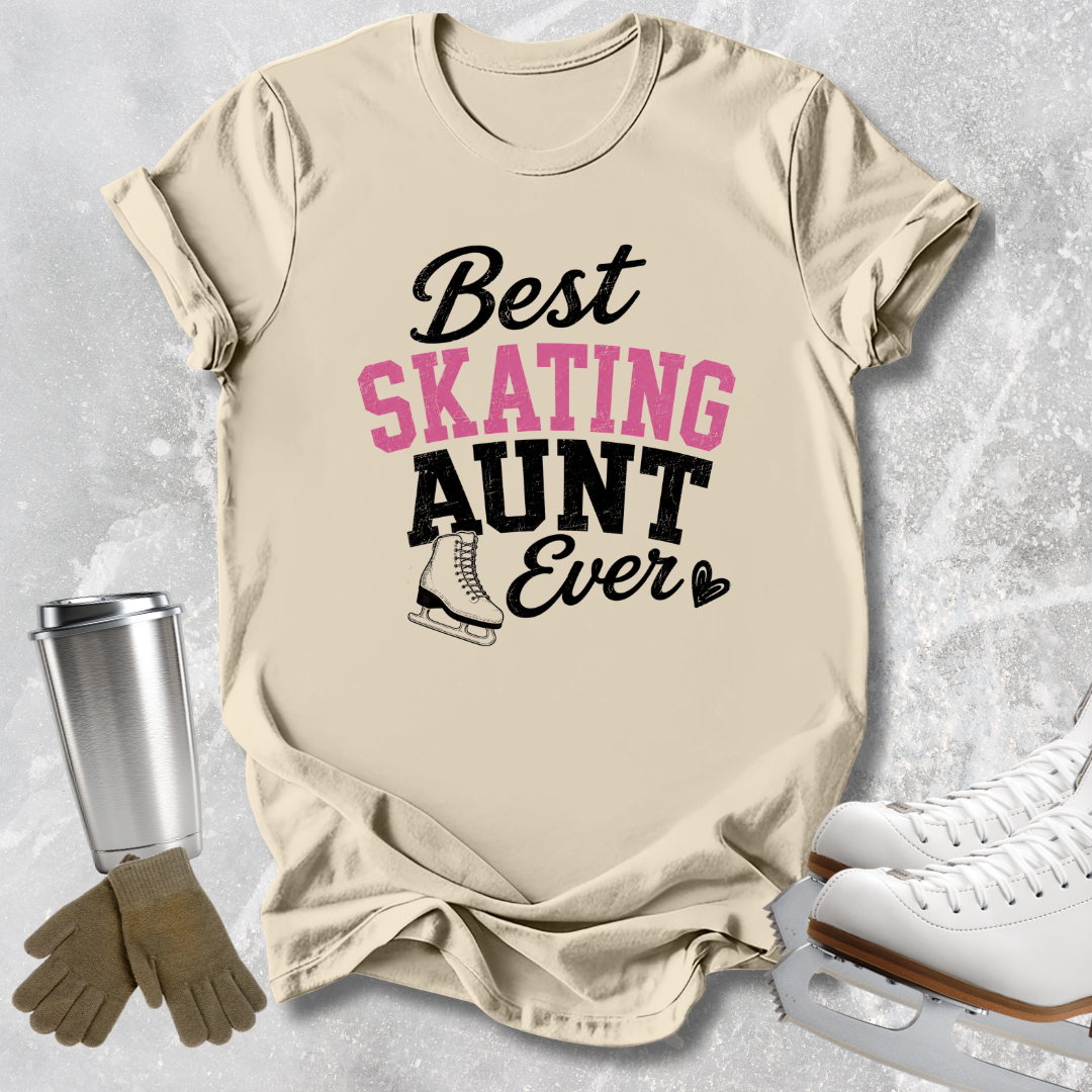 Beige graphic T-shirt laid flat on a light textured background, with the words 'Best SKATING AUNT Ever' in black and pink lettering and a small ice skate illustration. Surrounding props include white ice skates, gray gloves, and a stainless steel travel tumbler.