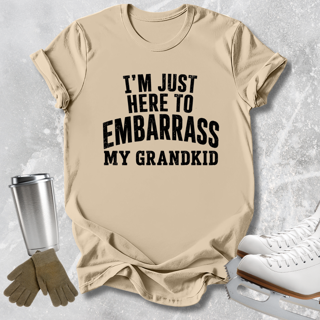 A beige graphic t-shirt with the text 'I'M JUST HERE TO EMBARRASS MY GRANDKID' in bold black letters, styled in a flat-lay with ice skates, gloves, and a tumbler on a light gray textured background.