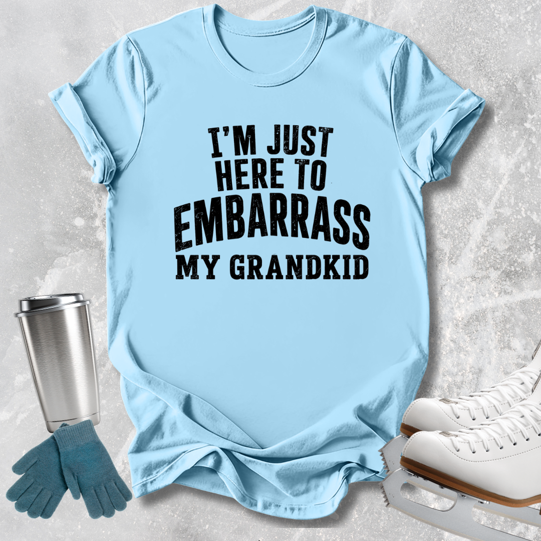 A light blue graphic t-shirt with bold black text reading 'I'M JUST HERE TO EMBARRASS MY GRANDKID', styled in a flat-lay with ice skates, gloves, and a tumbler on a textured gray background.
