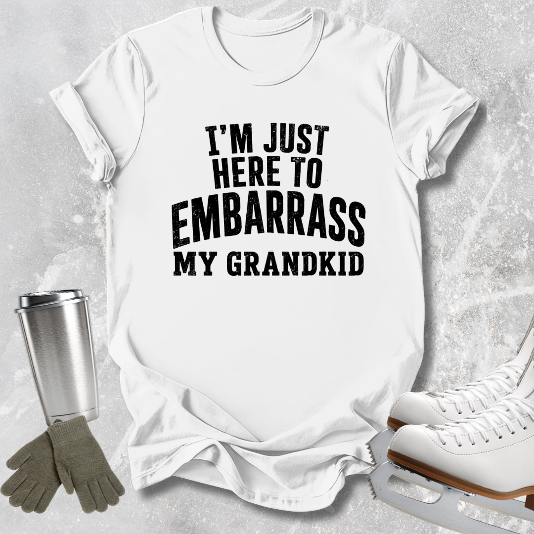 A white short-sleeve T-shirt with bold black text reading 'I'M JUST HERE TO EMBARRASS MY KID', styled in a flat lay with ice skates, gloves, and a metal cup on a light gray textured background.