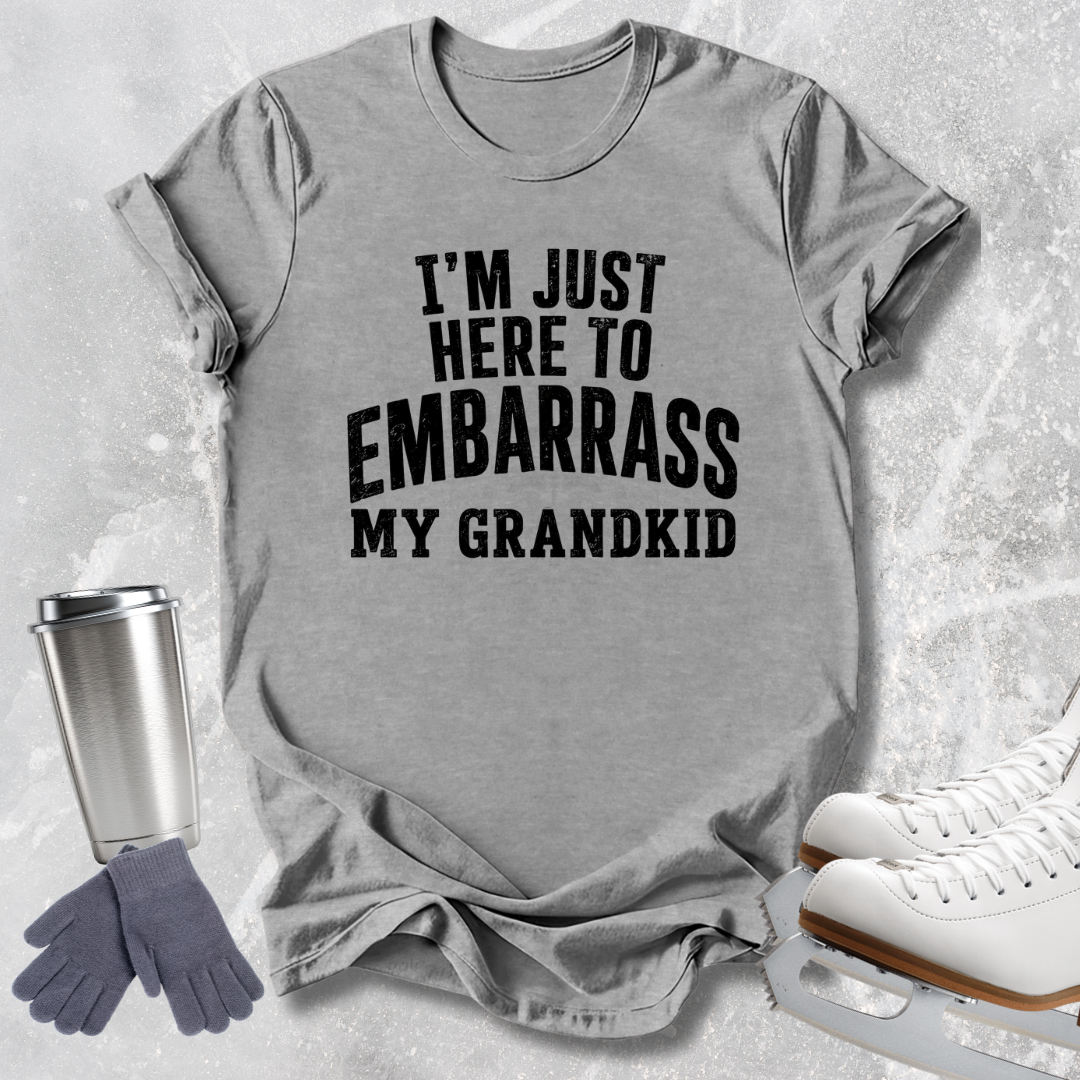 A heather gray graphic t-shirt with black text reading 'I'M JUST HERE TO EMBARRASS MY GRANDKID' is shown in a flat-lay arrangement with ice skates, a travel tumbler, and gloves on a light gray textured background.
