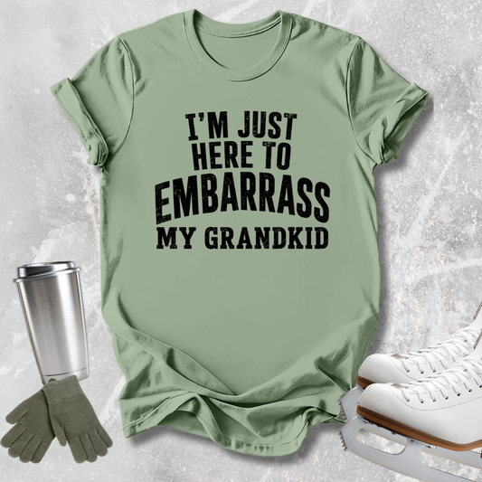 A sage green graphic t-shirt with the text 'I'M JUST HERE TO EMBARRASS MY GRANDKID' in black. The t-shirt is styled in a flat-lay arrangement on a light gray textured background, accompanied by a silver travel tumbler, gray gloves, and white ice skates.