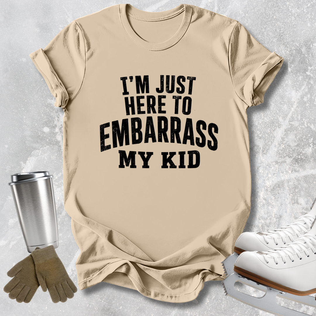 A beige graphic t-shirt with the text 'I'M JUST HERE TO EMBARRASS MY KID' in bold black letters, styled in a flat lay with ice skates, gloves, and a metal cup on a light gray textured background.