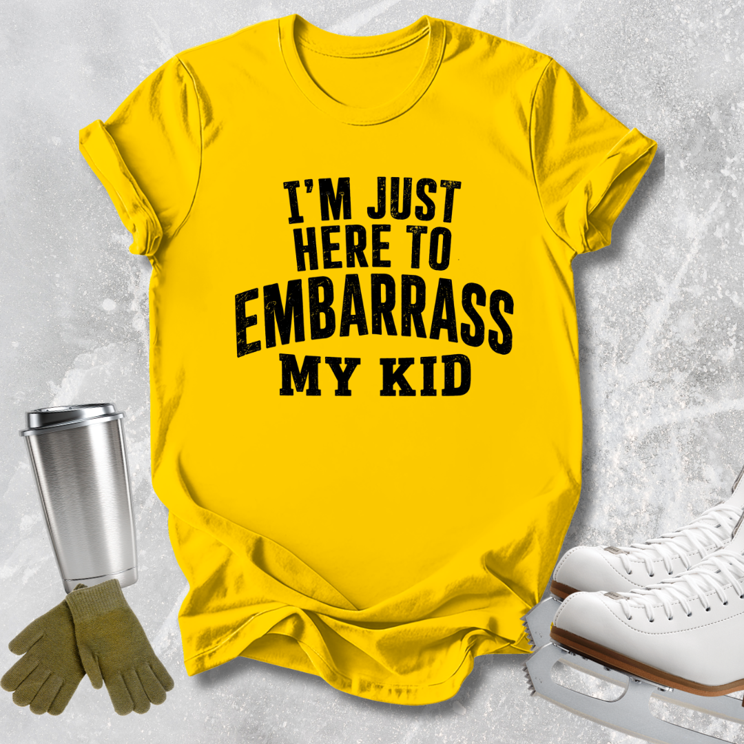 A bright yellow graphic t-shirt with bold black text reading 'I'M JUST HERE TO EMBARRASS MY KID', styled in a flat-lay arrangement with ice skates, gloves, and a metal cup on a light gray textured background.
