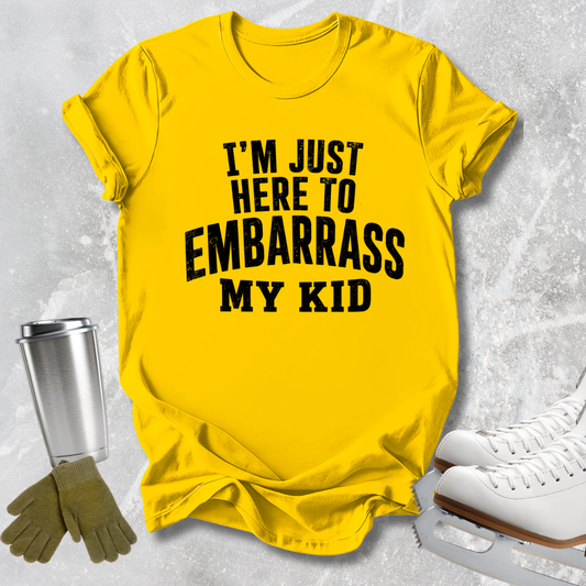 A bright yellow graphic t-shirt with bold black text reading 'I'M JUST HERE TO EMBARRASS MY KID', styled in a flat-lay arrangement with ice skates, gloves, and a metal cup on a light gray textured background.