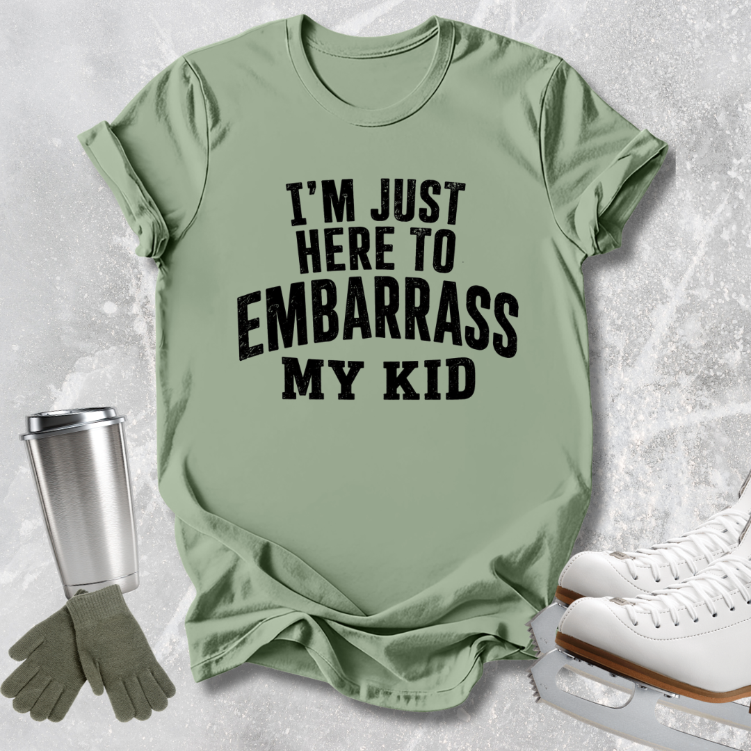 A sage green graphic t-shirt with the text 'I'M JUST HERE TO EMBARRASS MY KID' in bold black letters, styled in a flat-lay with ice skates, a tumbler, and gloves on a light gray textured background.