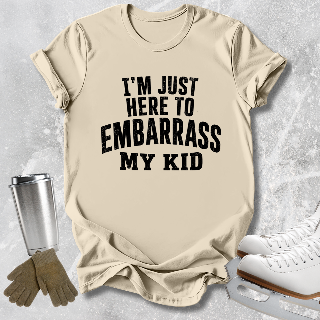 A beige graphic t-shirt with the text 'I'M JUST HERE TO EMBARRASS MY KID' in bold black letters, styled in a flat-lay with ice skates, gloves, and a metal cup on a light gray textured background.