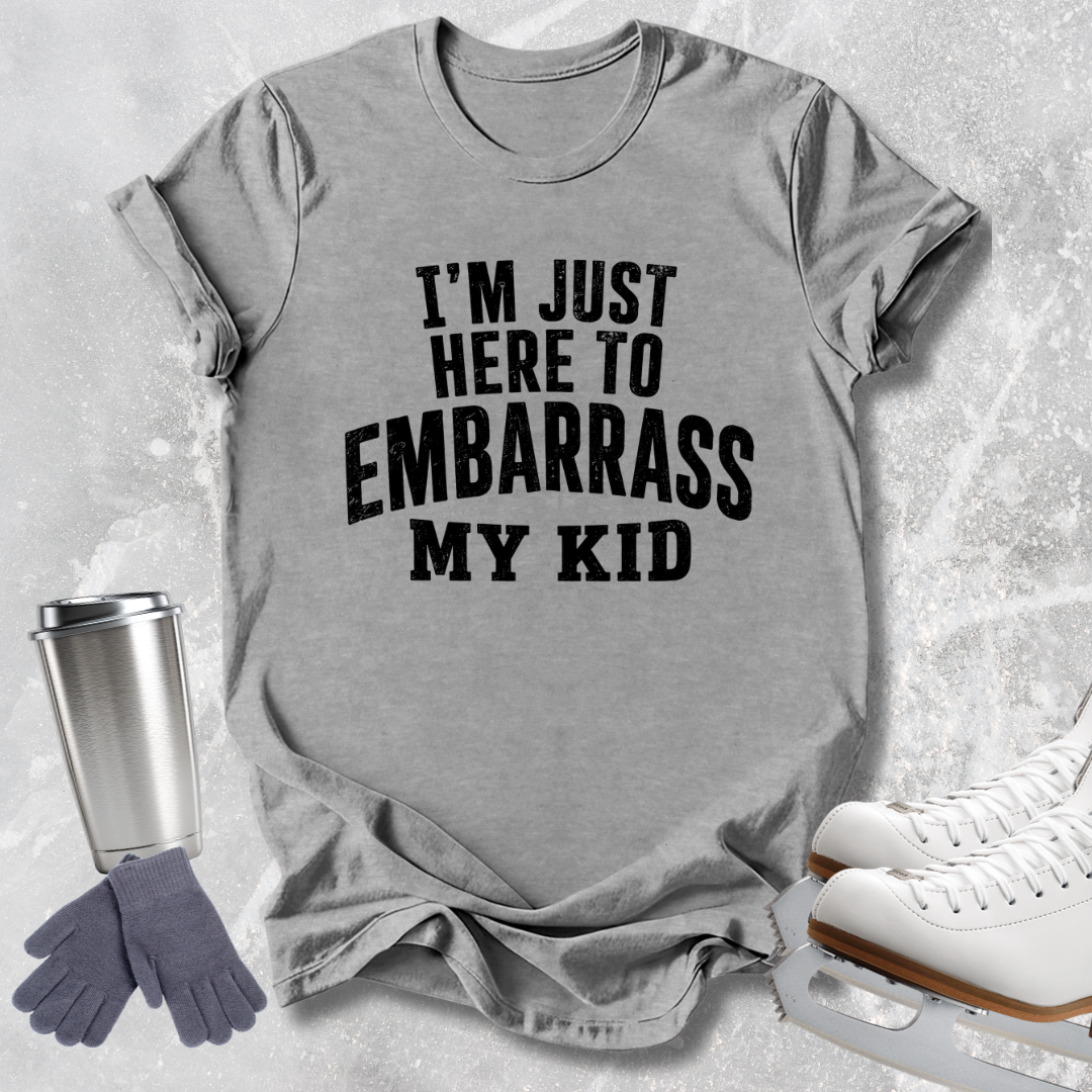 A heather gray graphic t-shirt with the text 'I'M JUST HERE TO EMBARRASS MY KID' in bold black letters, styled in a flat-lay with ice skates, a travel tumbler, and gloves on a light gray textured background.