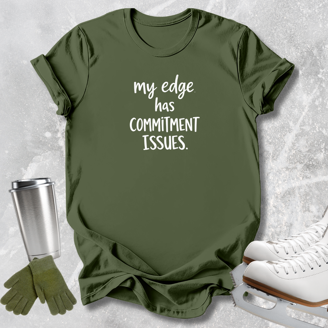 An olive green graphic t-shirt with the white printed text 'my edge has COMMITMENT ISSUES.' displayed in a flat-lay arrangement with ice skating shoes, a travel tumbler, and a pair of gloves on a light gray textured background.