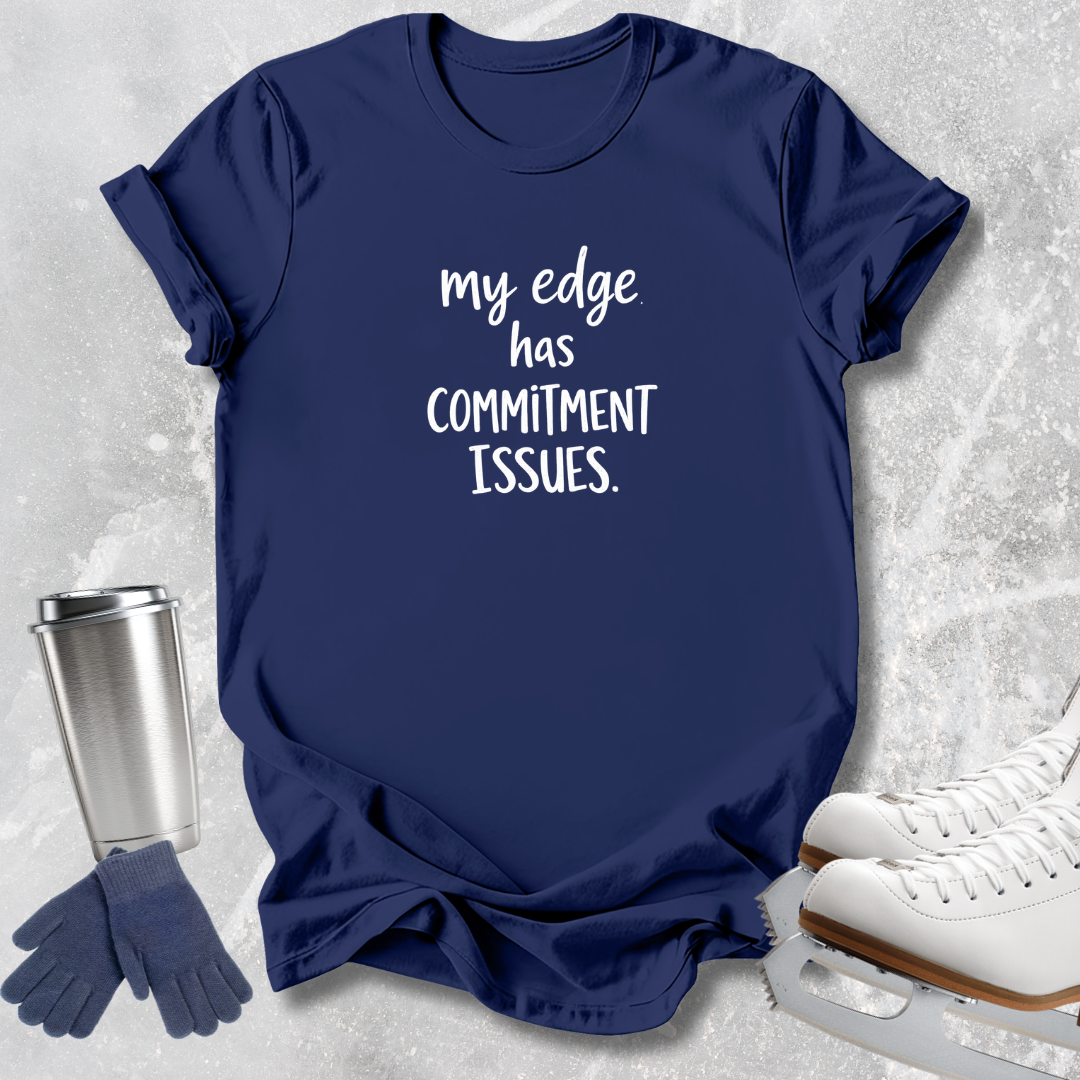 A navy blue graphic t-shirt with the white printed text 'my edge has COMMITMENT ISSUES.' displayed in a flat-lay arrangement with ice skate boots, gloves, and a travel tumbler on a light gray textured background.