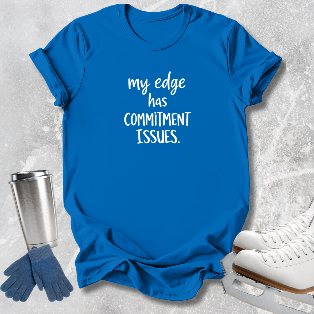 A bright blue short-sleeve t-shirt with white text reading 'my edge has COMMITMENT ISSUES.' laid flat on a light gray textured surface, styled with ice skates, gloves, and a metal travel tumbler.