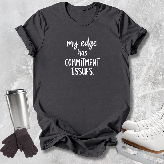 A dark charcoal gray graphic t-shirt with white text reading 'my edge has COMMITMENT ISSUES.' styled in a flat-lay arrangement with ice skates, gloves, and a metal tumbler on a light gray textured background.