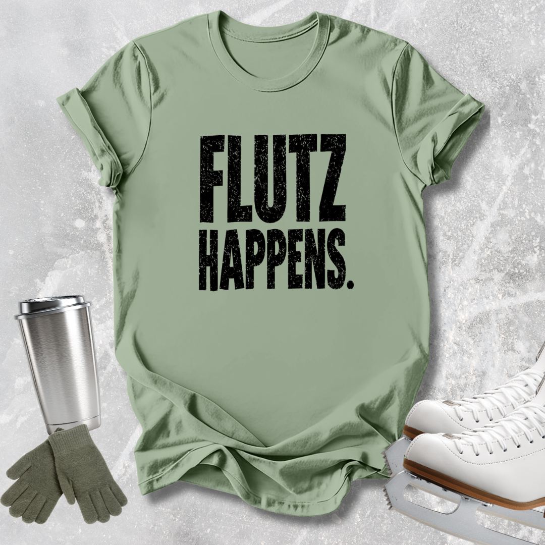 A sage green graphic t-shirt with the text 'FLUTZ HAPPENS.' printed in distressed black letters, styled in a flat-lay with ice skates, gloves, and a travel tumbler on a light gray textured background.