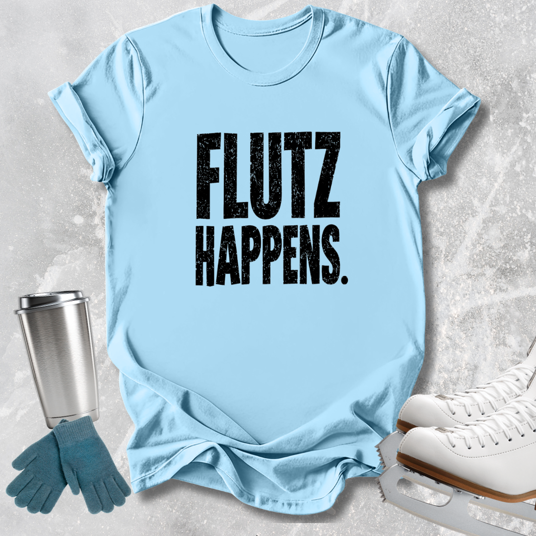 A light blue short-sleeve T-shirt with the phrase 'FLUTZ HAPPENS.' printed in large distressed black letters across the front, styled in a flat lay with ice skates, gloves, and a metal cup on a light gray textured background.