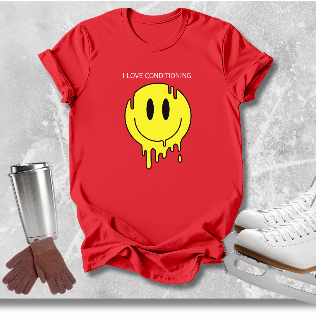 A red short-sleeve T-shirt laid flat on a light gray textured surface, featuring a yellow dripping smiley face graphic with the text 'I LOVE CONDITIONING' above it. Ice skates, gloves, and a metal cup are placed around the shirt as styled props.