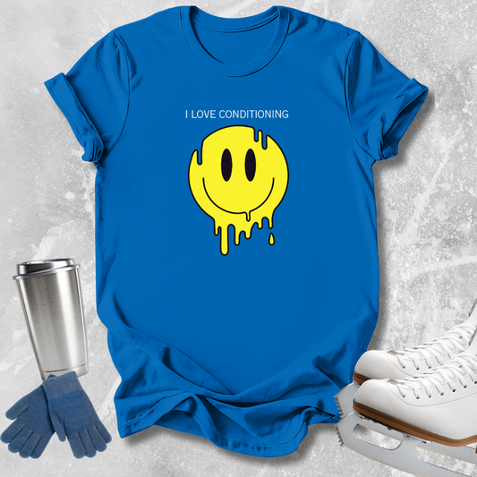 A bright blue short-sleeve T-shirt laid flat on a light textured surface, featuring a yellow dripping smiley face graphic with the text 'I LOVE CONDITIONING' above it. Blue gloves and white ice skates are placed around the shirt as styling props.