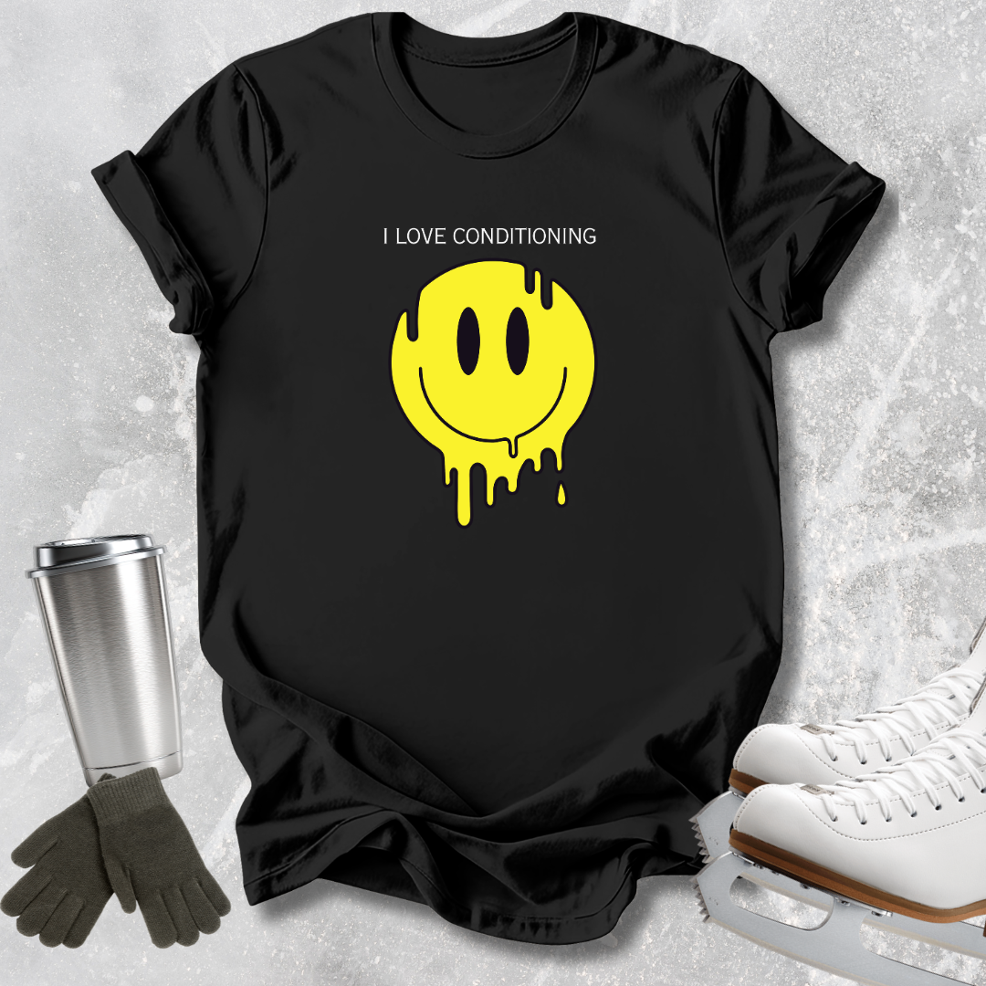 A black short-sleeve T-shirt laid flat on a light textured surface, featuring a yellow dripping smiley face graphic with the text 'I LOVE CONDITIONING' above it. A stainless steel cup, gloves, and white ice skates are arranged around the shirt as styling props.