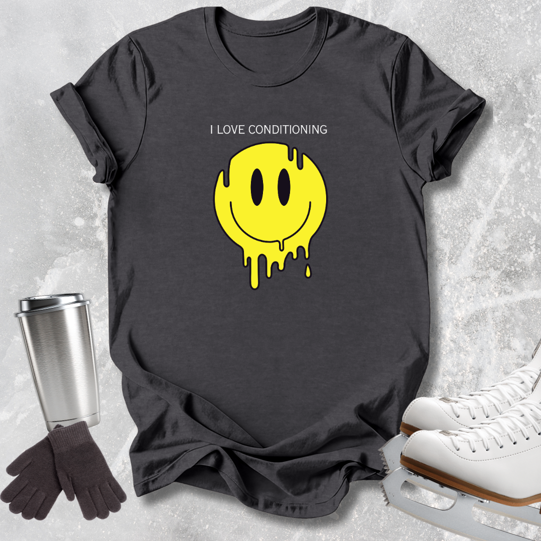 A dark charcoal gray graphic t-shirt laid flat on a light gray textured surface, featuring a yellow dripping smiley face and the text 'I LOVE CONDITIONING'. Surrounding props include a stainless steel travel mug, black gloves, and white ice skates.