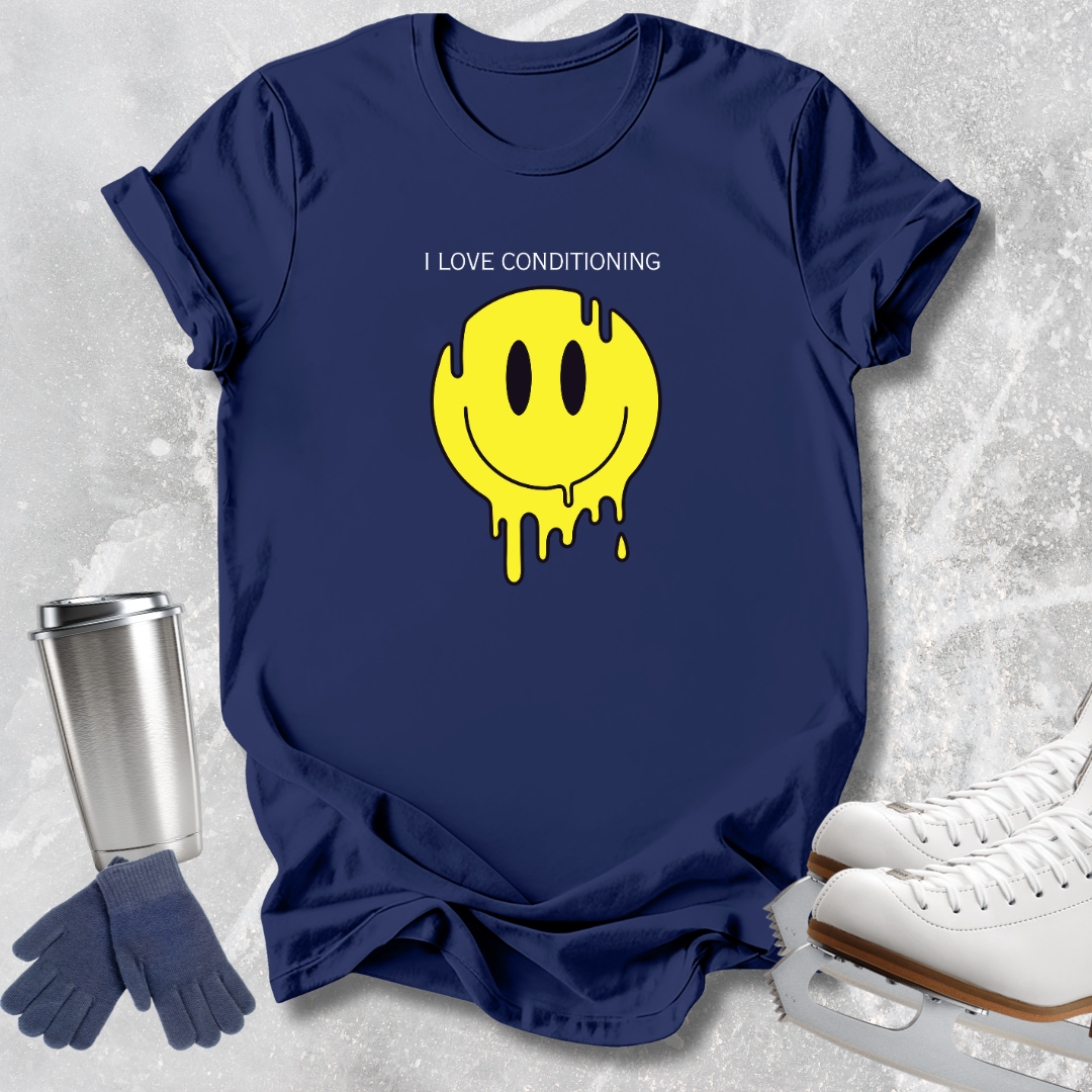 A navy blue short-sleeve T-shirt laid flat on a light gray textured surface, featuring the phrase 'I LOVE CONDITIONING' above a large yellow dripping smiley face graphic. A stainless steel travel tumbler, blue gloves, and white ice skates are arranged around the shirt as props.