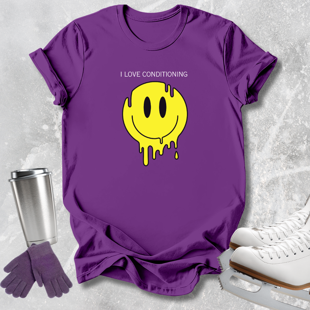 A purple short-sleeve T-shirt laid flat on a light gray textured surface, featuring a yellow dripping smiley face graphic with the text 'I LOVE CONDITIONING' above it. A stainless steel travel tumbler, purple gloves, and white ice skates are arranged around the shirt as styling props.
