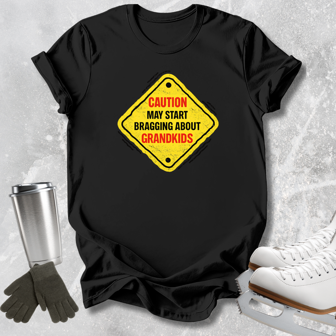 A black short-sleeve T-shirt laid flat on a light gray textured surface, featuring a yellow diamond-shaped warning sign graphic with red and black text that reads, 'CAUTION MAY START BRAGGING ABOUT GRANDKIDS.' A stainless steel tumbler, dark gloves, and white ice skates are arranged around the shirt as styling props.