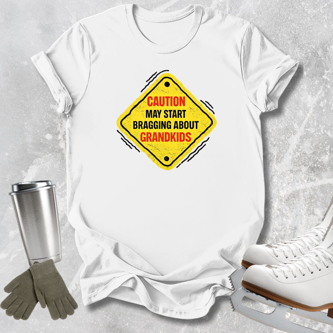 A white short-sleeve T-shirt laid flat on a light gray textured surface, featuring a yellow diamond-shaped warning sign graphic with the text 'CAUTION MAY START BRAGGING ABOUT GRANDKIDS' in red and black. A stainless steel tumbler, gray gloves, and white ice skates are placed around the shirt as styled props.