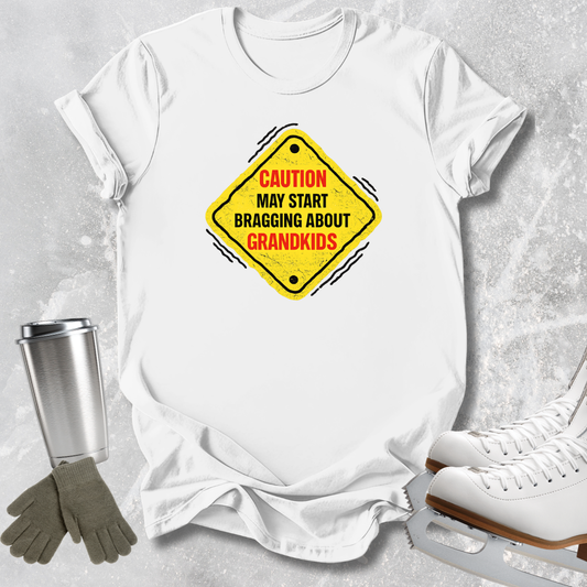 A white short-sleeve T-shirt laid flat on a light gray textured surface, featuring a yellow diamond-shaped warning sign graphic with the text 'CAUTION MAY START BRAGGING ABOUT GRANDKIDS' in red and black. A stainless steel tumbler, gray gloves, and white ice skates are placed around the shirt as styled props.