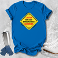 A bright blue short-sleeve T-shirt laid flat on a light gray textured surface, featuring a yellow diamond-shaped warning sign graphic with red and black text that reads, 'CAUTION MAY START BRAGGING ABOUT GRANDKIDS.' Blue gloves and white ice skates are placed around the shirt as styling props.