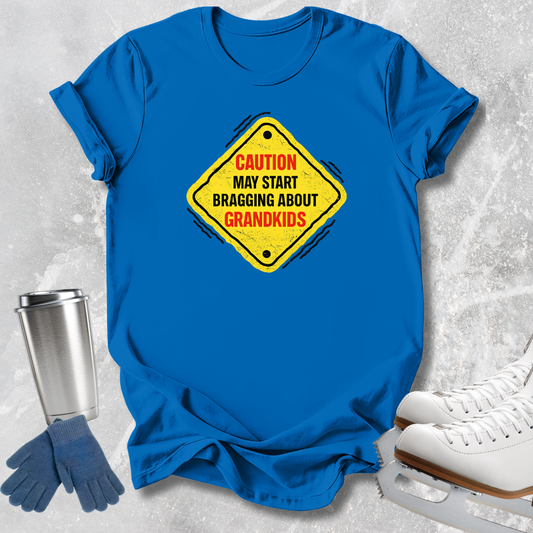 A bright blue short-sleeve T-shirt laid flat on a light gray textured surface, featuring a yellow diamond-shaped warning sign graphic with red and black text that reads, 'CAUTION MAY START BRAGGING ABOUT GRANDKIDS.' Blue gloves and white ice skates are placed around the shirt as styling props.