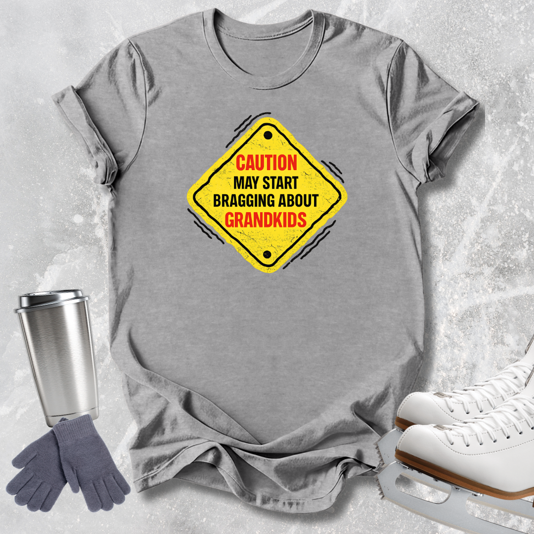A heather gray short-sleeve t-shirt with a yellow diamond-shaped warning sign graphic that reads 'CAUTION MAY START BRAGGING ABOUT GRANDKIDS' in red and black text. The shirt is styled in a flat-lay arrangement on a light gray textured background, accompanied by a stainless steel travel tumbler, gray gloves, and white ice skates.