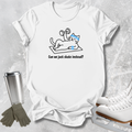 A white graphic t-shirt laid flat on a light gray textured surface, featuring a cartoon cat wearing a blue headband and lifting a dumbbell with the text 'Can we just skate instead?'. Ice skates, gloves, and a metal tumbler are styled around the shirt.