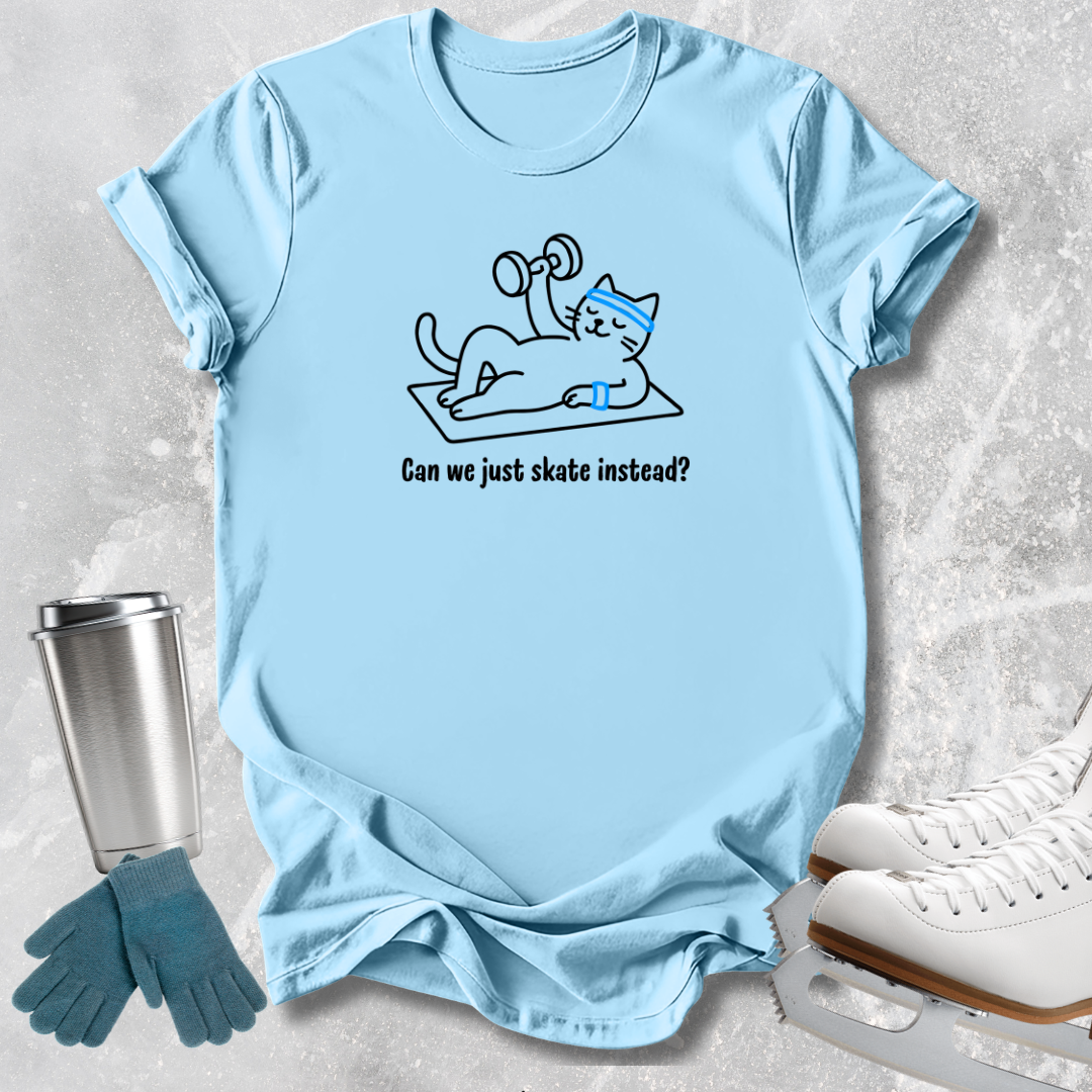A light blue graphic t-shirt laid flat on a textured gray background, featuring a cartoon cat lifting a dumbbell with the text 'Can we just skate instead?'. Ice skates, gloves, and a metal tumbler are styled around the shirt.