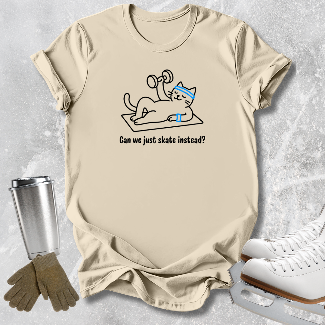 A beige graphic t-shirt laid flat on a light gray textured surface, featuring a cartoon cat doing pushups with dumbbells and the text 'Can we just skate instead?'. White ice skates, a silver tumbler, and tan gloves are styled around the shirt.
