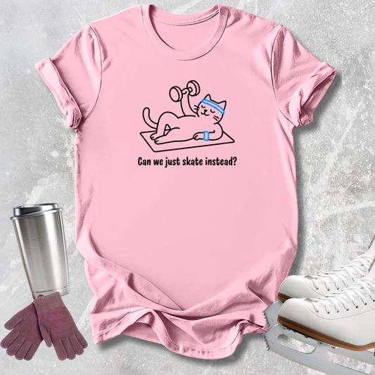 A pink short-sleeve graphic T-shirt laid flat on a light gray textured surface, with a cartoon cat lifting weights and the text 'Can we just skate instead?' on the front. A stainless steel tumbler, purple gloves, and white ice skates are arranged around the shirt as styling props.