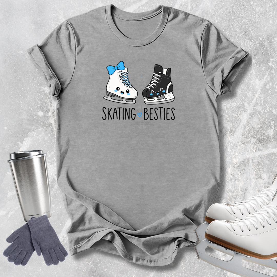 A heather gray short-sleeve T-shirt laid flat on a light stone-like surface, featuring a graphic of two cartoon ice skates with the text 'SKATING BESTIES.' A stainless steel travel mug, gray gloves, and white ice skates appear as styled props around the shirt.