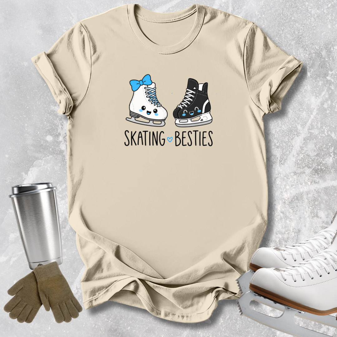 A beige graphic t-shirt laid flat on a textured gray background, featuring a cartoon illustration of a white ice skate with a blue bow and a black ice skate, with the text 'SKATING BESTIES' underneath. A stainless steel travel mug, a pair of brown gloves, and white ice skates are styled around the t-shirt.