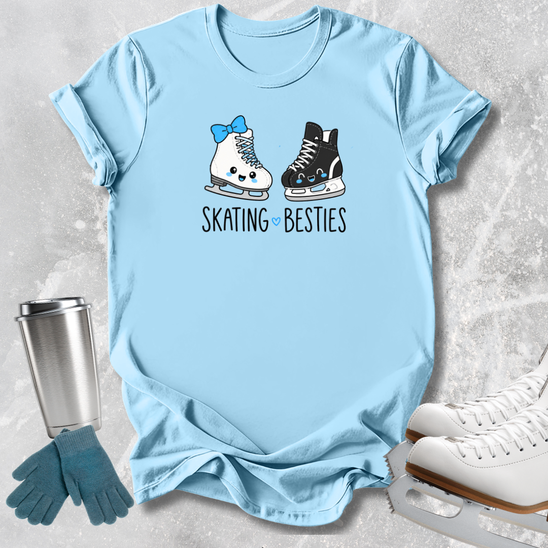 A light blue graphic t-shirt laid flat on a textured gray background, featuring a cute illustration of a blue bow on one ice skate and the text 'SKATING BESTIES' below it. Styled flat-lay props include a stainless steel tumbler, teal gloves, and white ice skates.