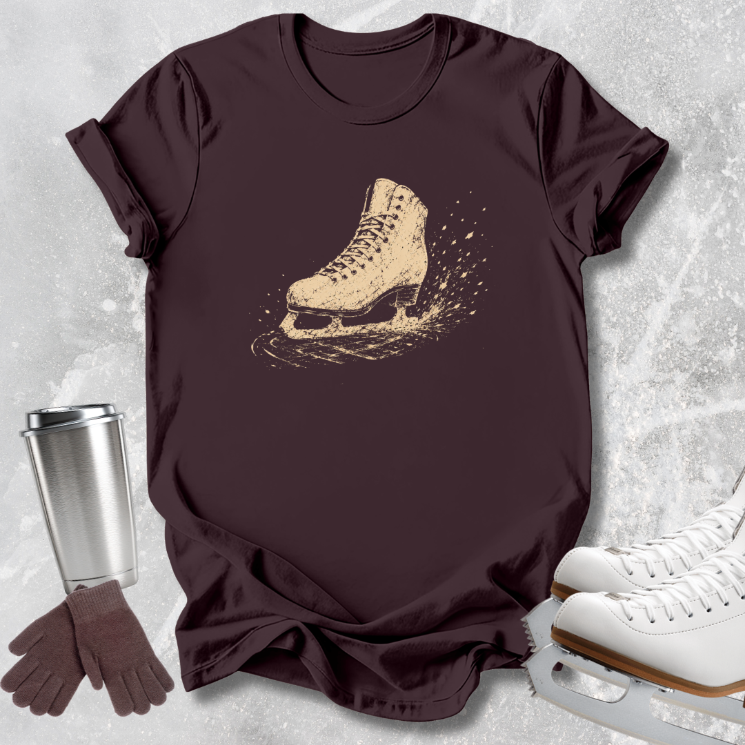 A dark brown short-sleeve t-shirt with a beige graphic of an ice skate, styled in a flat lay with white ice skates, a silver travel mug, and a pair of brown gloves on a light gray textured background.