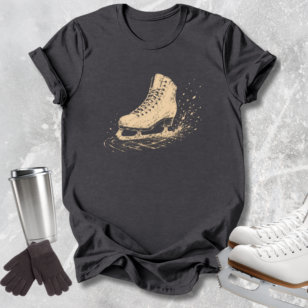 A dark charcoal gray t-shirt featuring a vintage-style graphic of a beige ice skate boot with a splash effect, styled in a flat-lay with white ice skates, a silver tumbler, and a pair of black gloves on a light gray textured background.