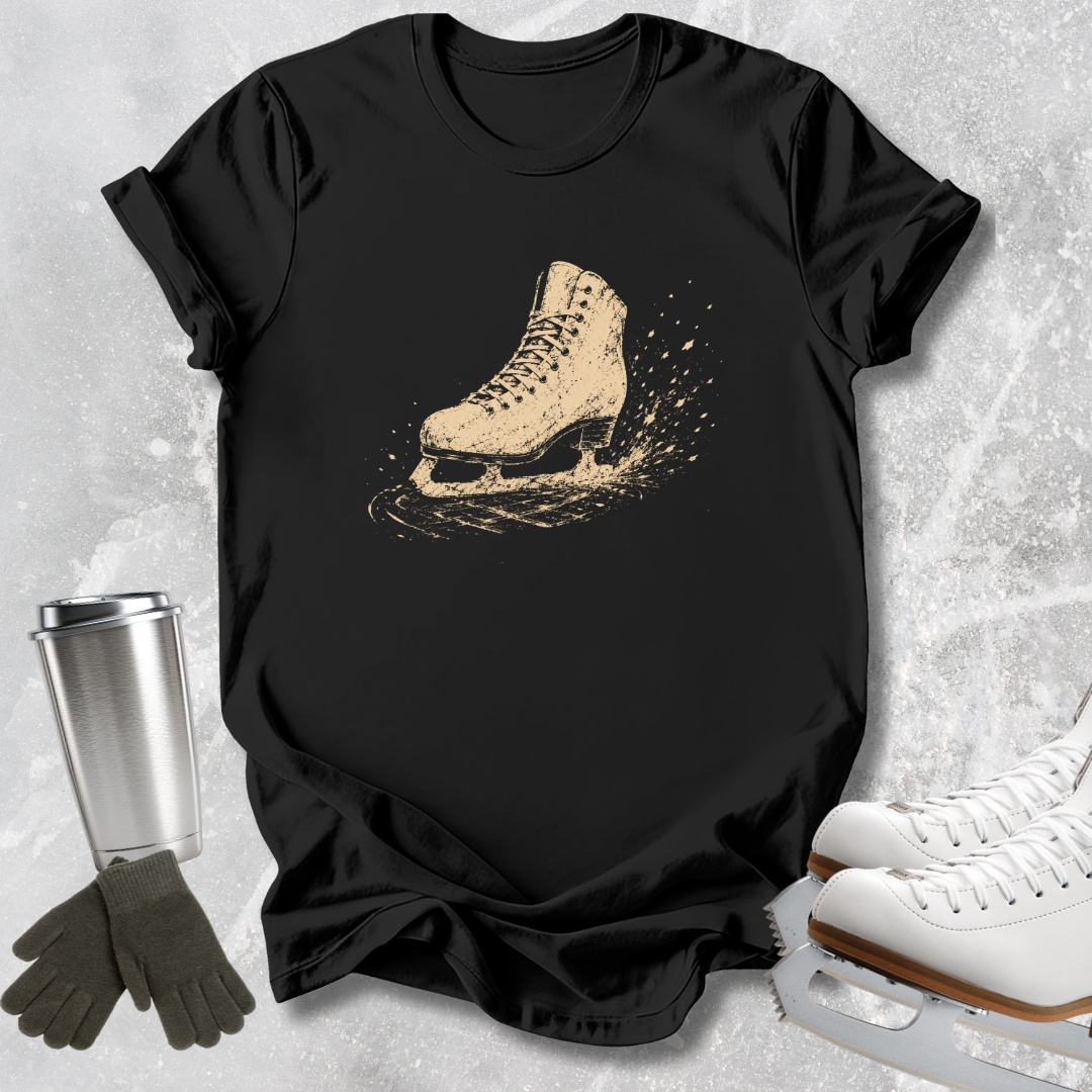 A black short-sleeve T-shirt laid flat on a light textured surface, featuring a distressed beige ice skate graphic on the chest. Surrounding props include a silver travel mug, dark gloves, and white ice skates.