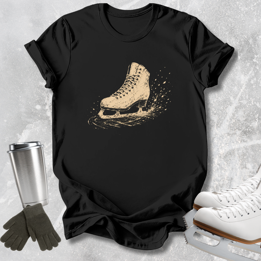 A black short-sleeve T-shirt laid flat on a light textured surface, featuring a distressed beige ice skate graphic on the chest. Surrounding props include a silver travel mug, dark gloves, and white ice skates.