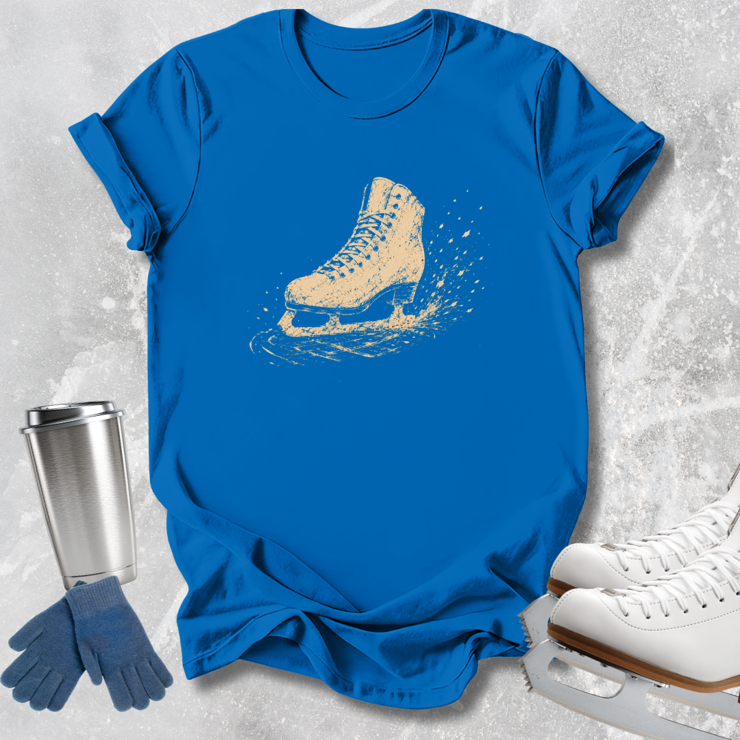 A bright blue short-sleeve T-shirt laid flat on a light gray textured surface, featuring a distressed beige ice skate graphic on the chest. Blue gloves and a silver tumbler are placed beside the shirt, with white ice skates partially visible at the lower right.