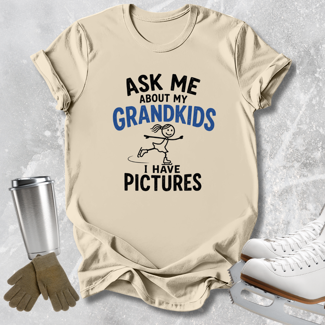 A beige graphic t-shirt laid flat on a light gray textured background, surrounded by ice skating props including white ice skates, a metal tumbler, and gloves. The shirt features a black and blue graphic that reads 'ASK ME ABOUT MY GRANDKIDS I HAVE PICTURES' with a stick-figure skater illustration.