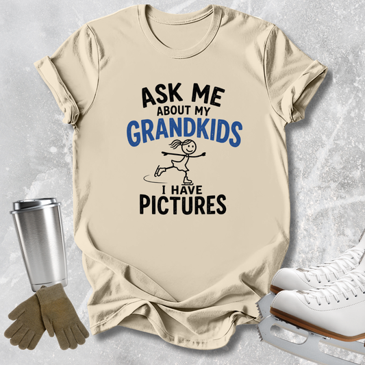 A beige graphic t-shirt laid flat on a light gray textured background, surrounded by ice skating props including white ice skates, a metal tumbler, and gloves. The shirt features a black and blue graphic that reads 'ASK ME ABOUT MY GRANDKIDS I HAVE PICTURES' with a stick-figure skater illustration.