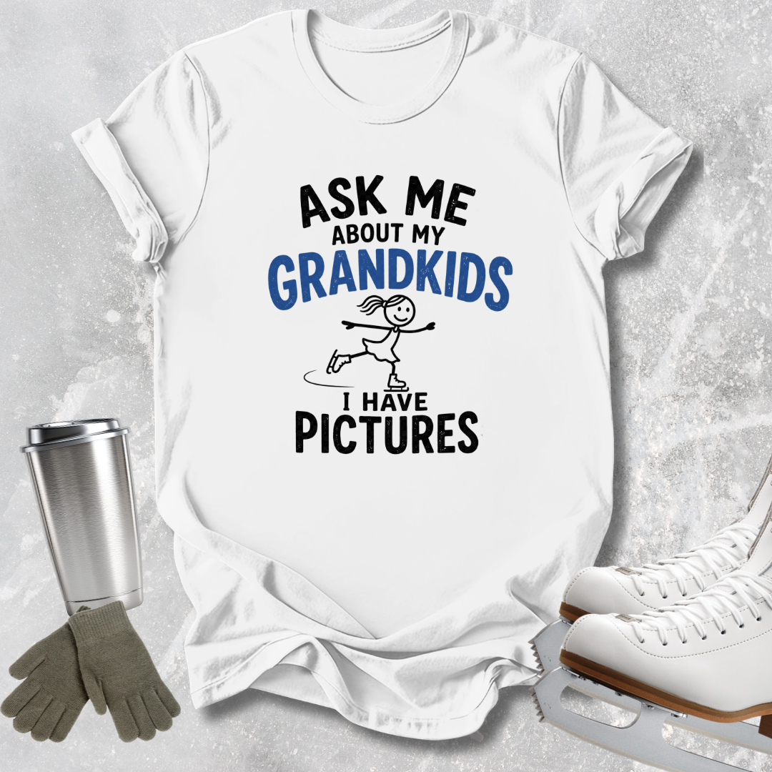 A white graphic t-shirt laid flat on a light gray textured background, with printed text reading 'ASK ME ABOUT MY GRANDKIDS I HAVE PICTURES' and a small skater illustration. Surrounding props include white ice skates, gray gloves, and a silver tumbler.