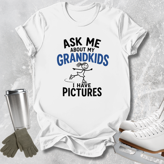 A white graphic t-shirt laid flat on a light gray textured background, with printed text reading 'ASK ME ABOUT MY GRANDKIDS I HAVE PICTURES' and a small skater illustration. Surrounding props include white ice skates, gray gloves, and a silver tumbler.