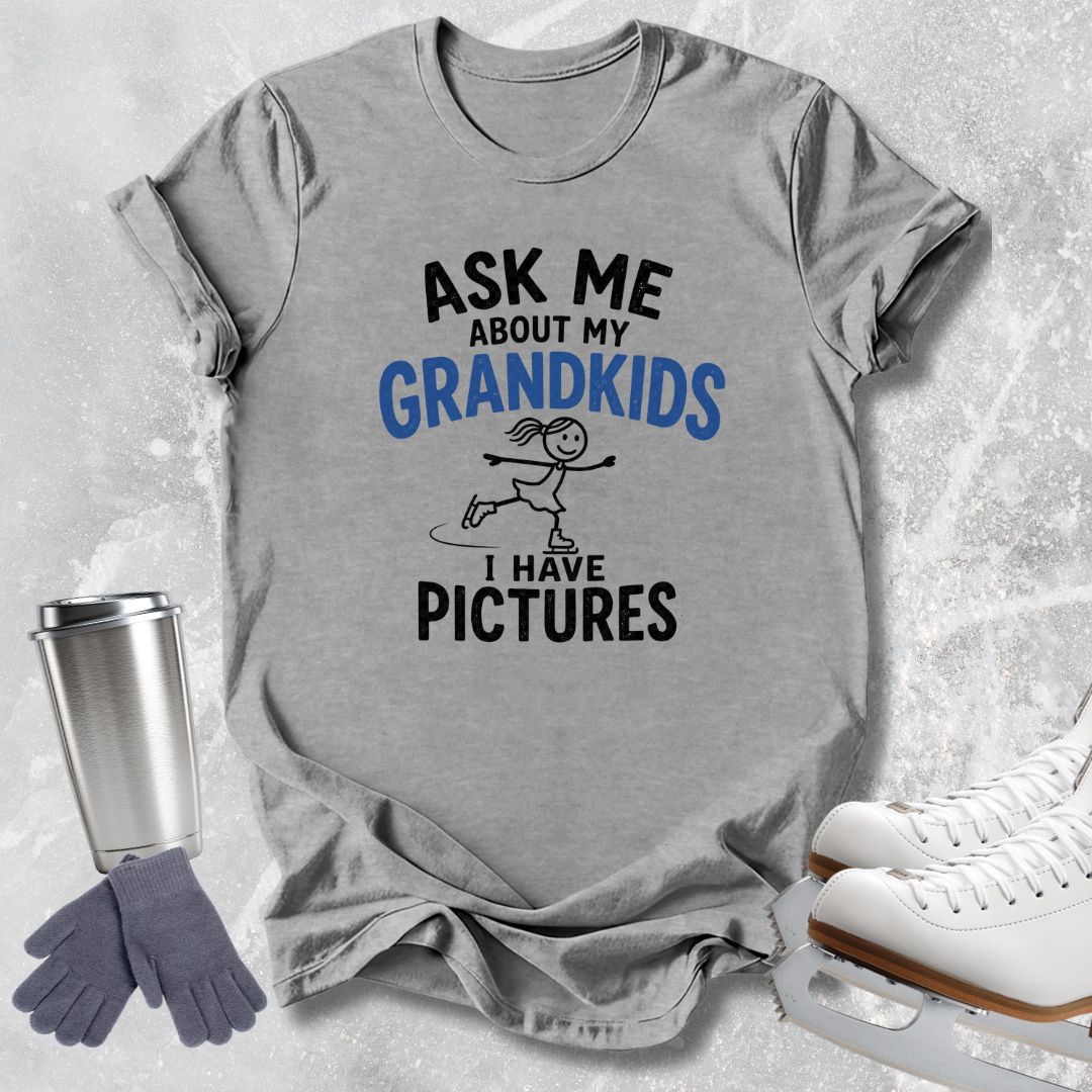 A heather gray graphic t-shirt laid flat on a light gray textured background, featuring the text 'ASK ME ABOUT MY GRANDKIDS I HAVE PICTURES' in black and blue lettering with a simple line drawing of a girl ice skating. Surrounding props include a stainless steel travel tumbler, a pair of gray gloves, and white ice skates.